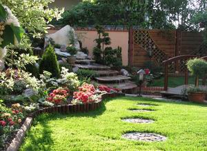 Ogród z kaskadą wodną, ścieżką z kamiennych płyt na trawniku i ozdobnymi roślinami. W tle drewniany płot i mały mostek.